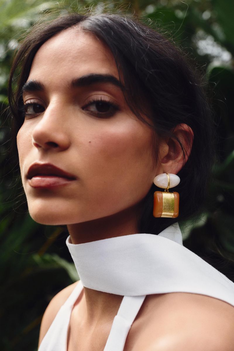 Close-up portrait of a woman wearing statement drop earrings and a white top against a lush green background.