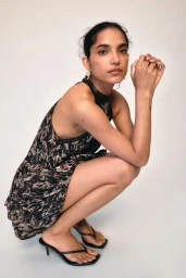 Model crouching against a neutral white background wearing a patterned black halter dress and heeled sandals