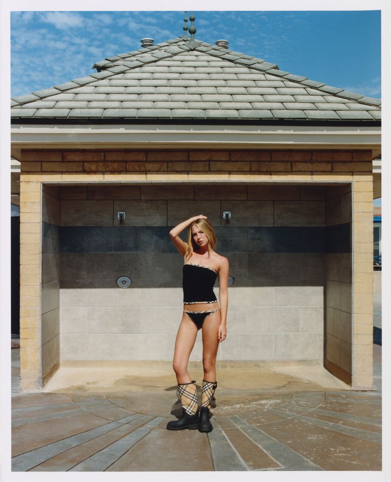 Model Lila Moss standing outdoors wearing a black bikini and patterned boots in front of a tiled structure.