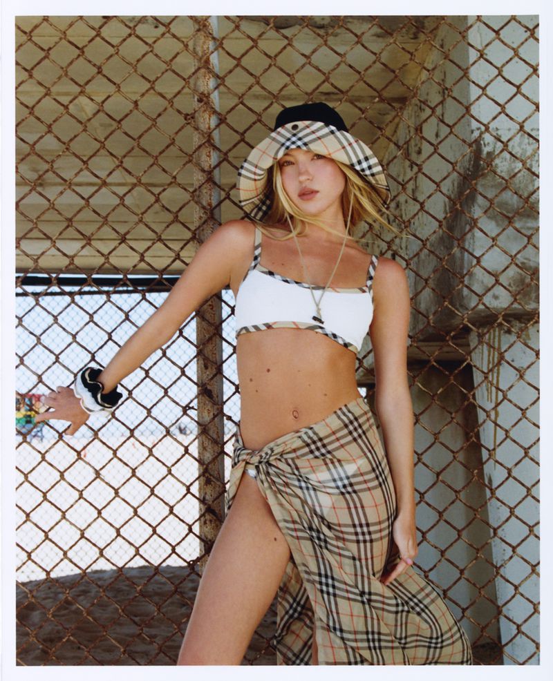 Model Lila Moss wearing plaid bucket hat and matching sarong against chain-link fence for Who What Wear