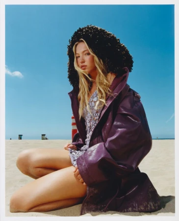 A blonde woman in a purple coat with a black fur hood sits on a sandy beach with blue sky, wearing a patterned dress in natural light.