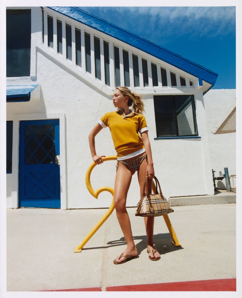 Model Lila Moss wearing a yellow polo shirt and holding a plaid handbag outdoors against a white building