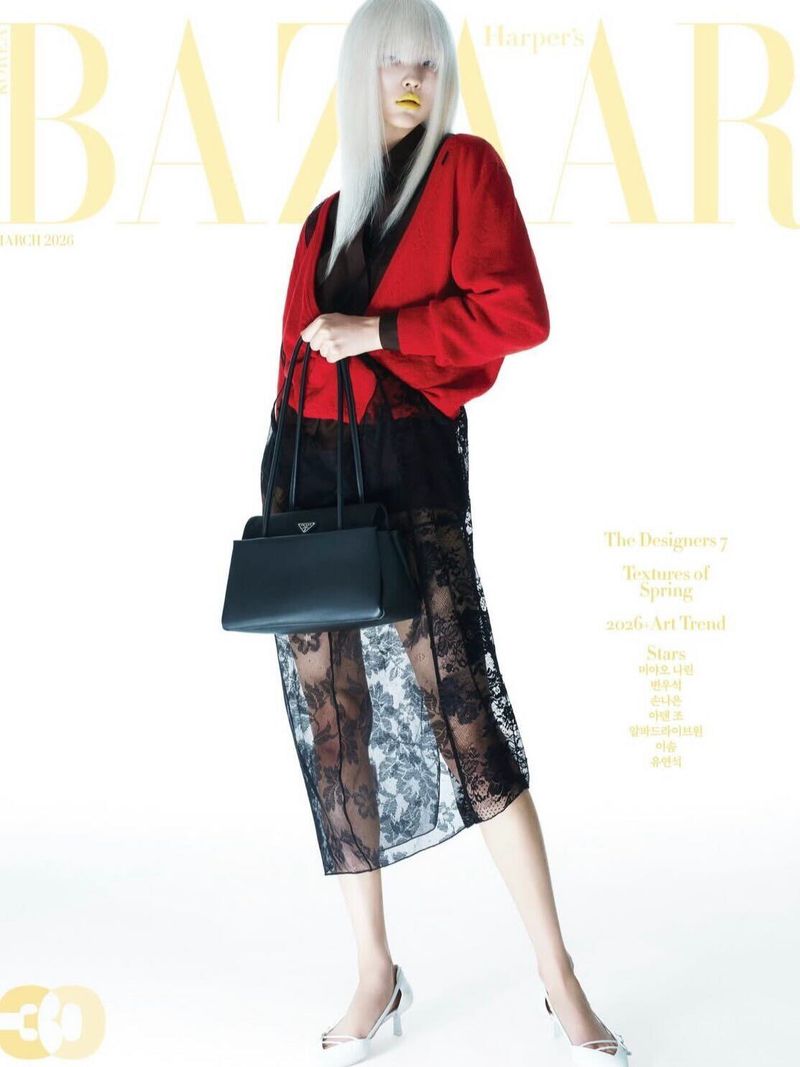 Fashion editorial shot of a woman with a platinum wig, red cardigan, black lace dress, white heels, and black Prada handbag against white background.