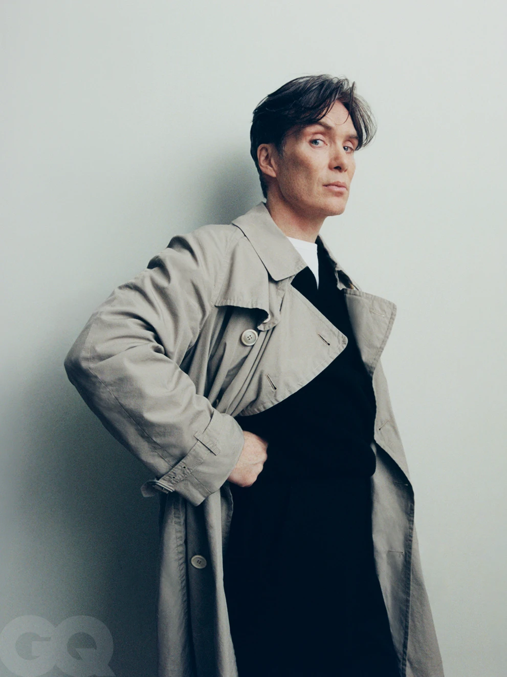 Actor Cillian Murphy wearing a beige trench coat over a black shirt against a plain grey studio backdrop.