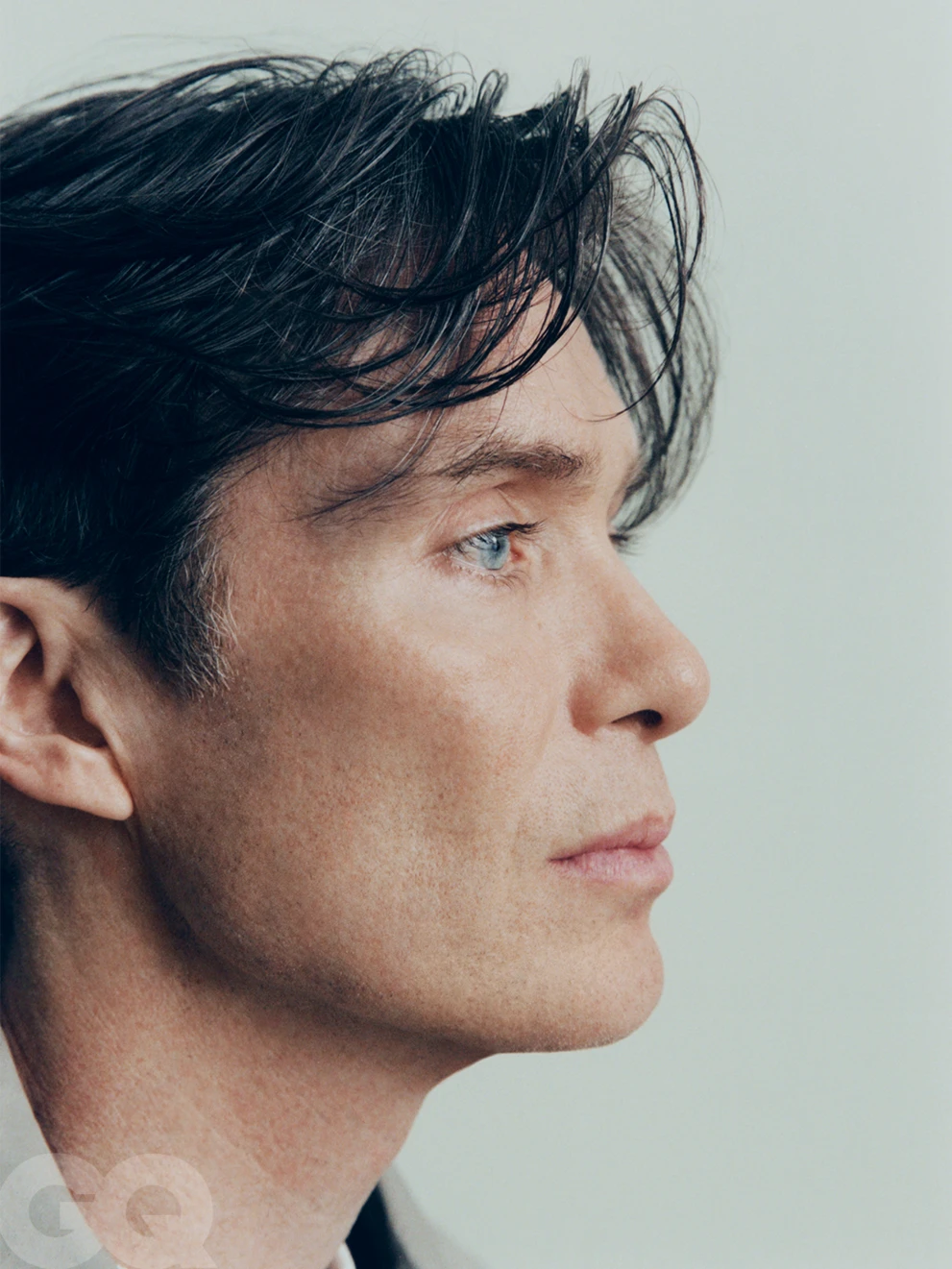 Close-up profile portrait of actor Cillian Murphy with textured dark hair against a pale neutral backdrop.