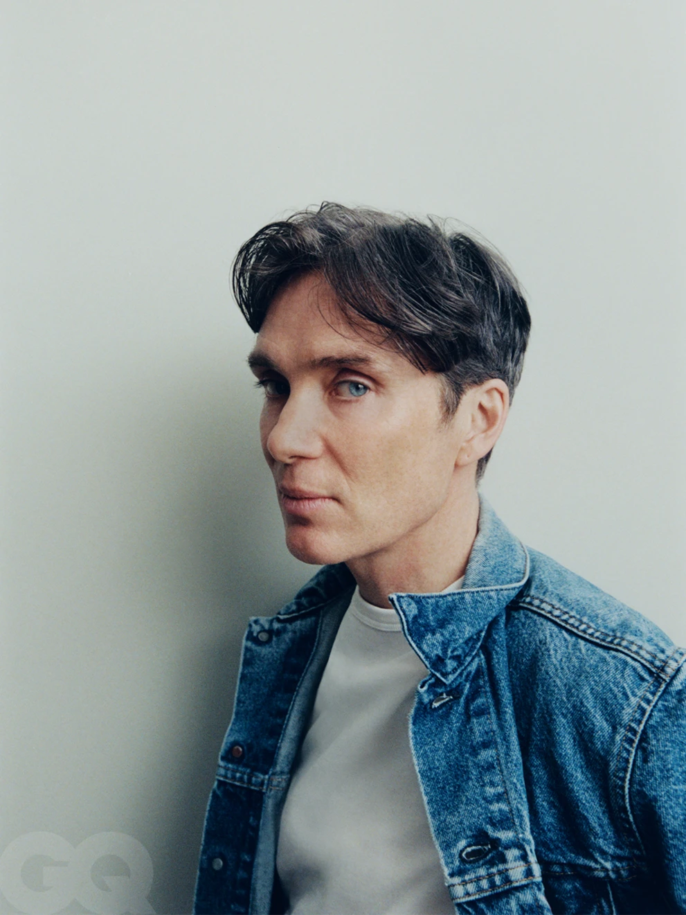 Portrait of actor Cillian Murphy wearing a blue denim jacket over a white t shirt against a neutral grey wall