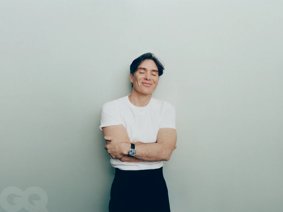 Cillian Murphy smiling with eyes closed in a white t-shirt and black trousers against a pale seamless studio background.