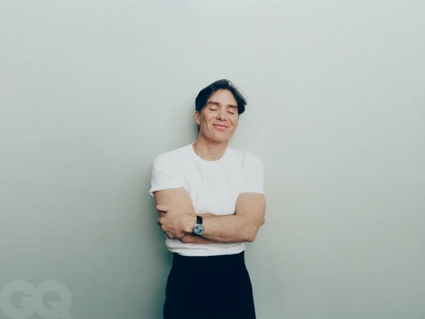 Cillian Murphy smiling with eyes closed in a white t-shirt and black trousers against a pale seamless studio background.