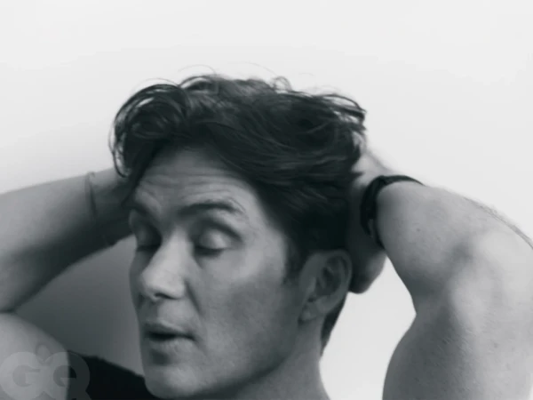 Monochrome portrait of Cillian Murphy with hands behind his head against a plain white background