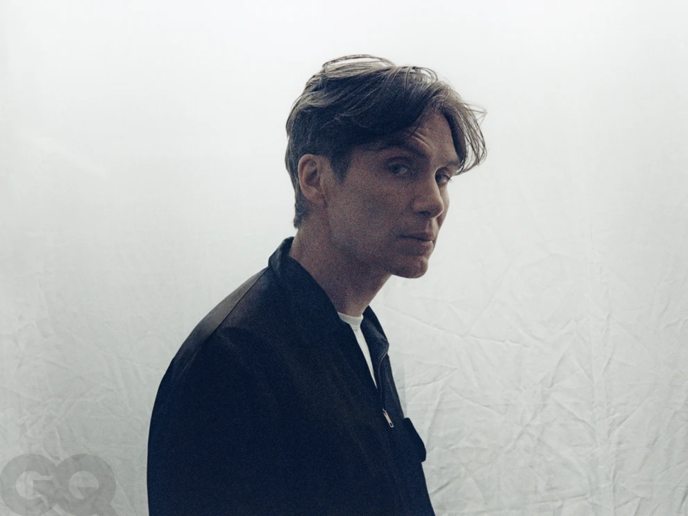 Actor Cillian Murphy looking over his shoulder in a black zip-up jacket against a creased white backdrop.