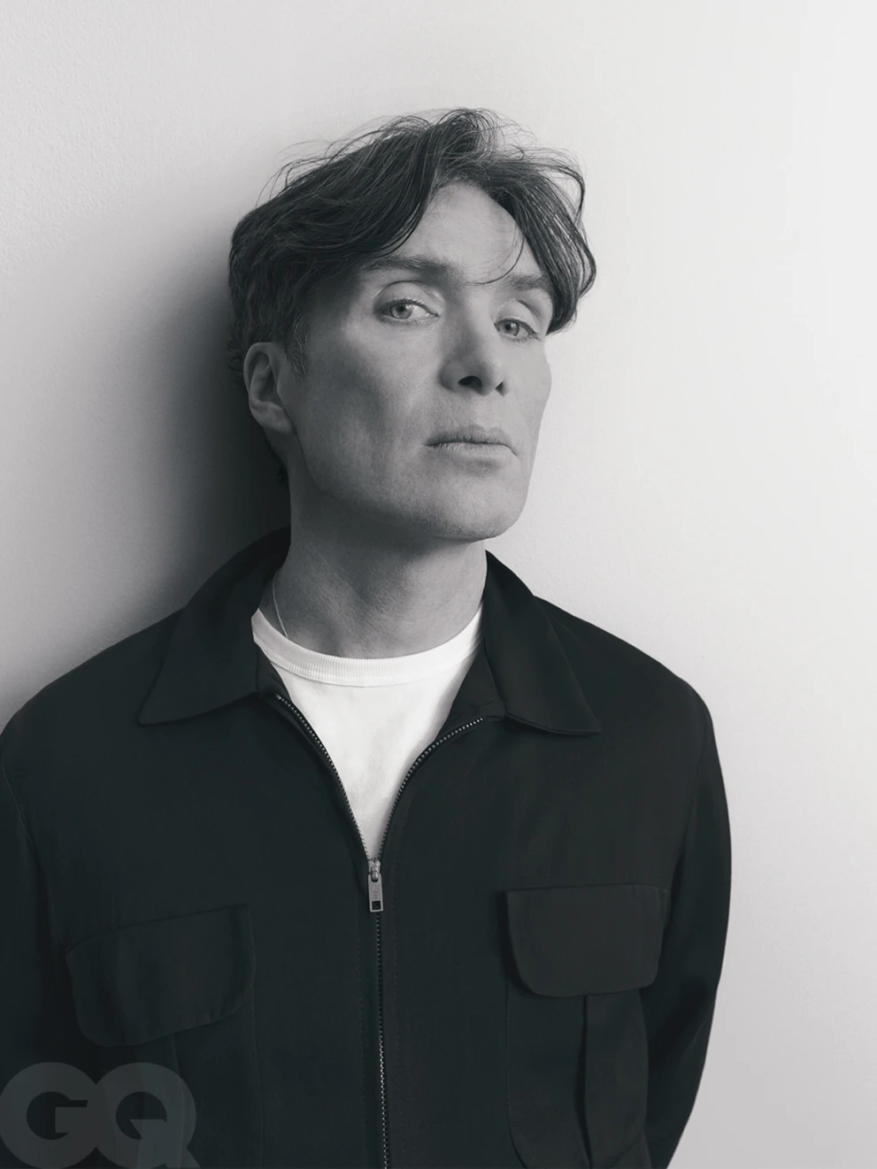 Portrait of Cillian Murphy wearing a black zip jacket over a white t-shirt against a plain white wall background