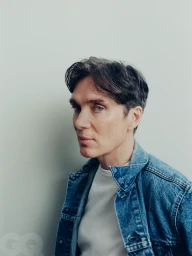 Portrait of actor Cillian Murphy wearing a blue denim jacket over a white t shirt against a neutral grey wall