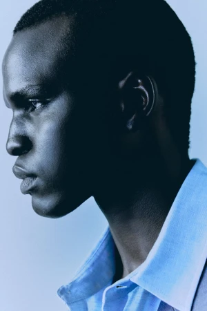 Close-up of an adult African male in a light blue collared shirt against a light blue background in a portrait-style fashion shot.