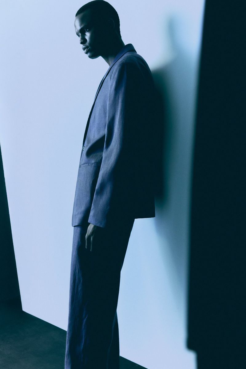 Portrait of a Black male model in a blue suit leaning against a wall in a minimalist studio setting with creative directional lighting.