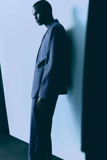 Portrait of a Black male model in a blue suit leaning against a wall in a minimalist studio setting with creative directional lighting.