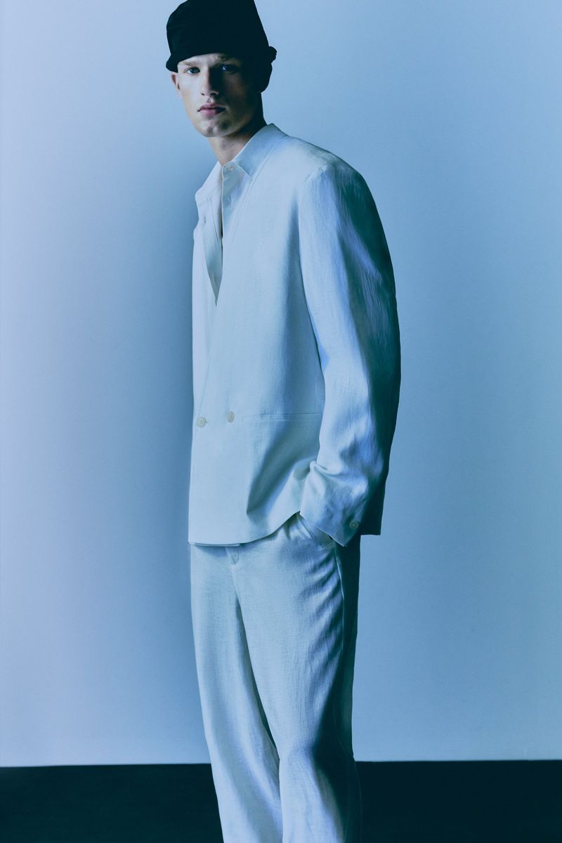 A man in a light-colored suit and black hat stands against a light blue background in a fashion shot, looking into the camera.