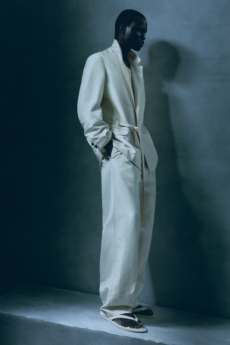 Adult Black man in a light suit and sandals, posed in a photo studio with soft, directional lighting, promoting a fashionable and relaxed style.