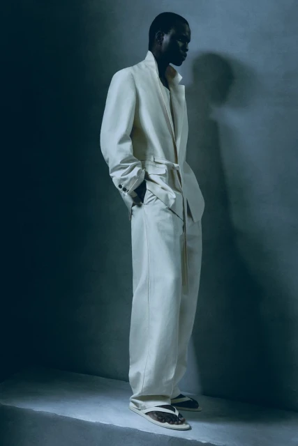 Adult Black man in a light suit and sandals, posed in a photo studio with soft, directional lighting, promoting a fashionable and relaxed style.