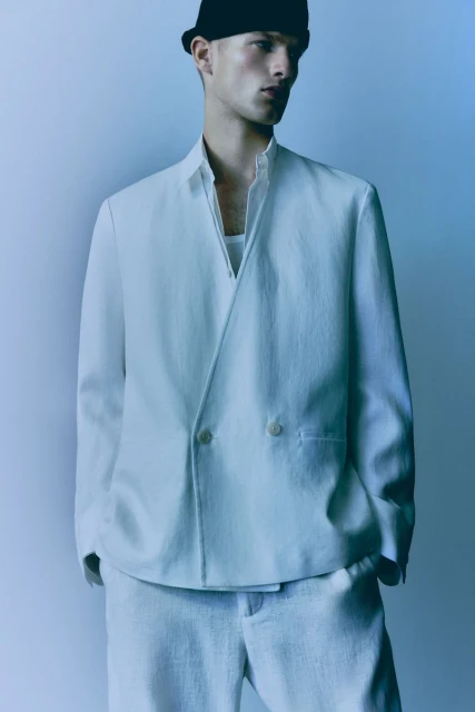 Man in light suit and black hat, wearing an open-collared white shirt. Soft studio lighting on a minimalist backdrop. Fashion forward style.