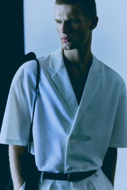 Portrait of adult male model in white suit jacket and pants with shoulder bag against a white background in a fashion photography studio.