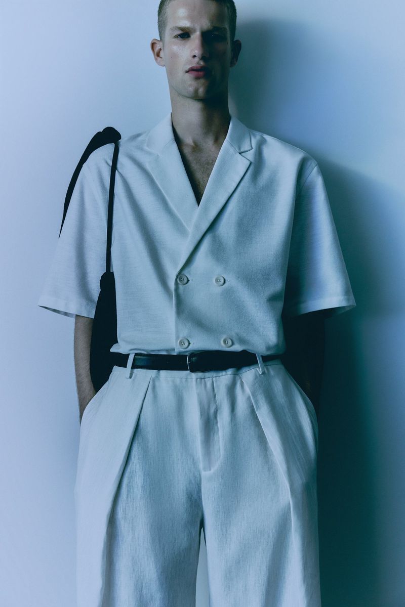 Studio shot of a male model in a light-colored, double-breasted short-sleeve shirt and wide-leg pants, wearing a black belt and shoulder bag.