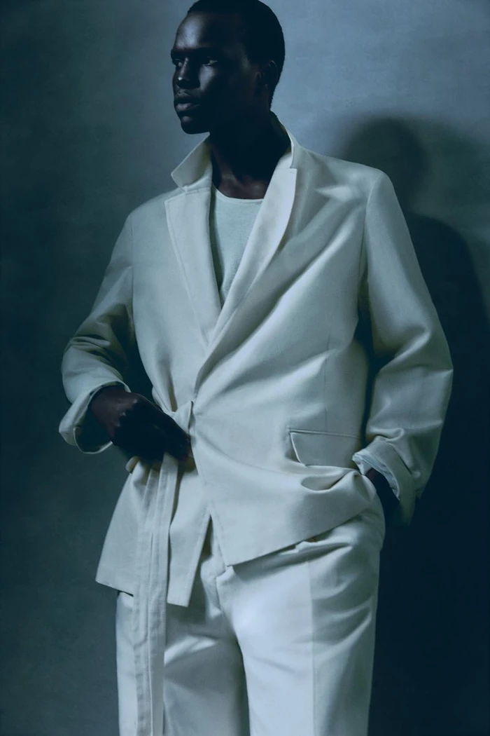 Model wearing a white suit jacket with a tie belt against a textured grey studio background