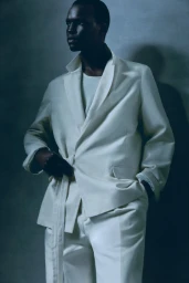 Model wearing a white suit jacket with a tie belt against a textured grey studio background