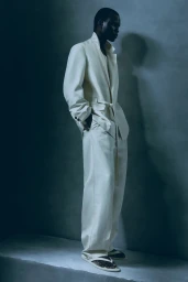 Black male model wearing a relaxed off-white belted suit and sandals standing against a cool grey studio backdrop.