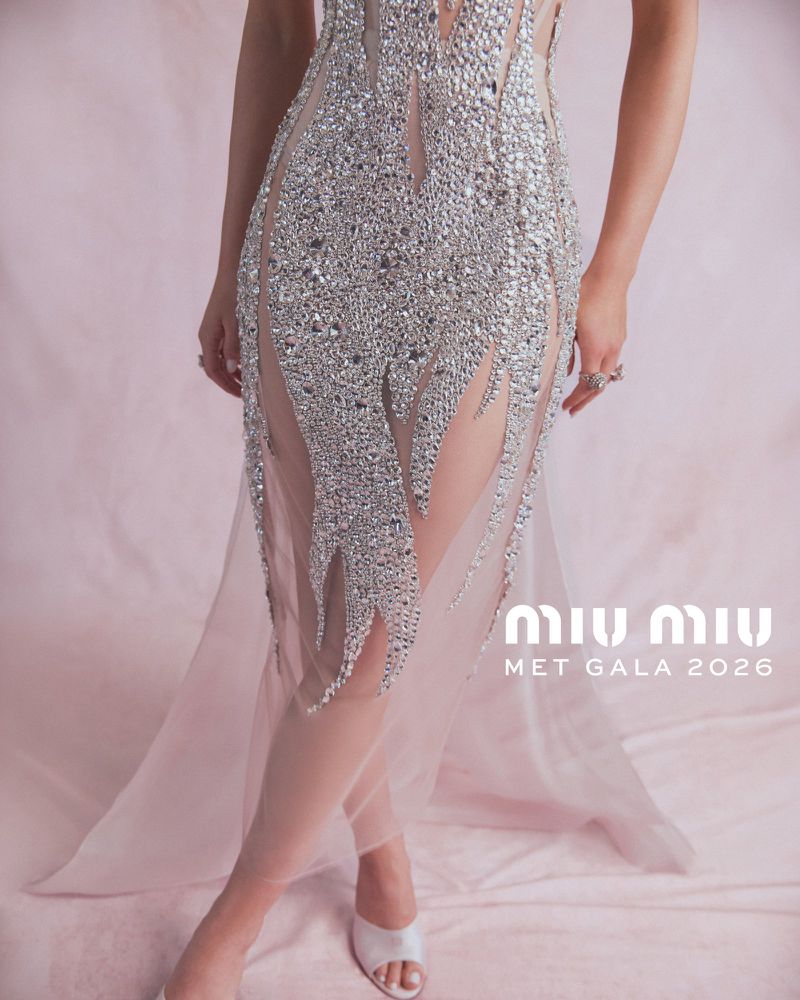 A fashion shot of an adult female model wearing a jeweled gown with sheer panels on a pink background, wearing white heels.