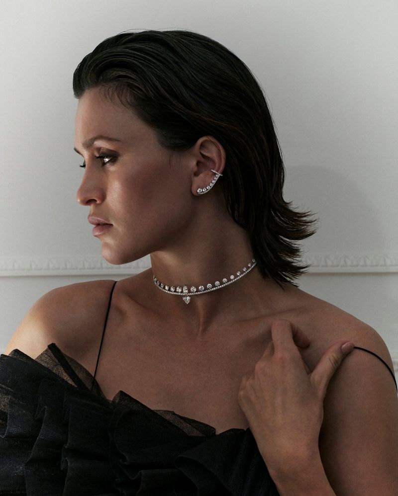 Close-up profile portrait of a woman wearing a diamond choker necklace and matching ear cuff against a plain white wall