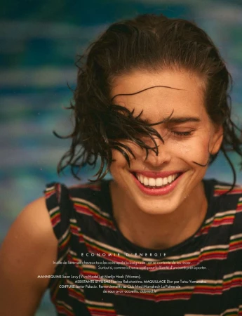 Close-up of a smiling woman with wet short dark hair covering her eye, wearing a multi-striped sleeveless top by a pool.