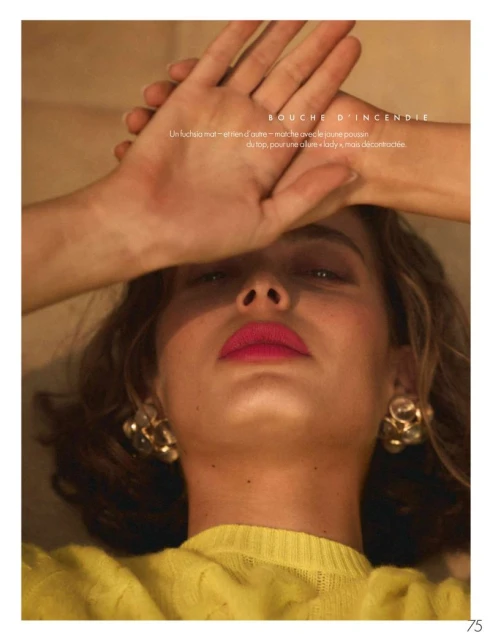 Close-up portrait of a woman with bright pink lipstick and gold earrings, hands raised above her forehead, wearing a yellow sweater.