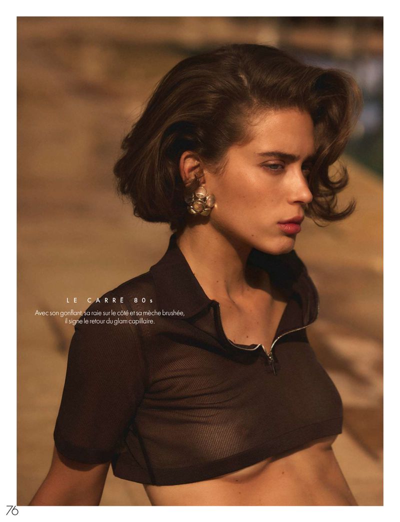 Fashion shot of a brunette woman with short hair, wearing a brown see-through top, and gold earrings, posing against a brown background.