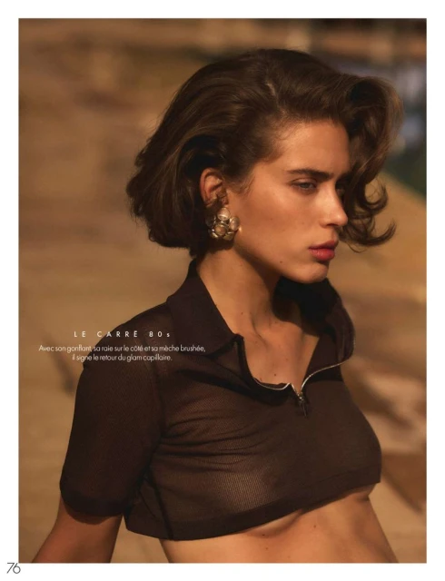 Fashion shot of a brunette woman with short hair, wearing a brown see-through top, and gold earrings, posing against a brown background.