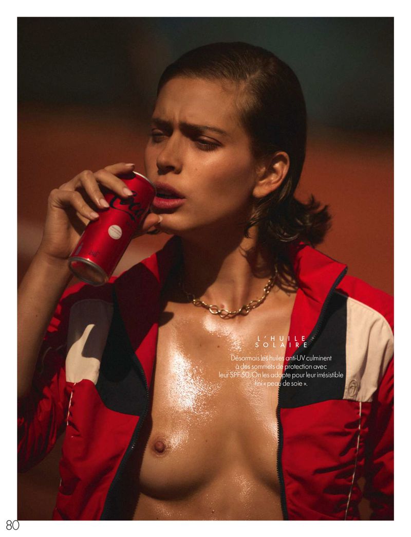 Woman in a red jacket drinks from a can in a close-up shot, showcasing her wet skin and gold necklace against a blurred background.