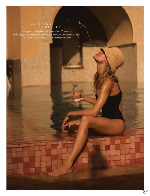 Woman in black swimsuit and straw hat sits by pool holding a bottle of water, catching water droplets on her face in the sunlight.
