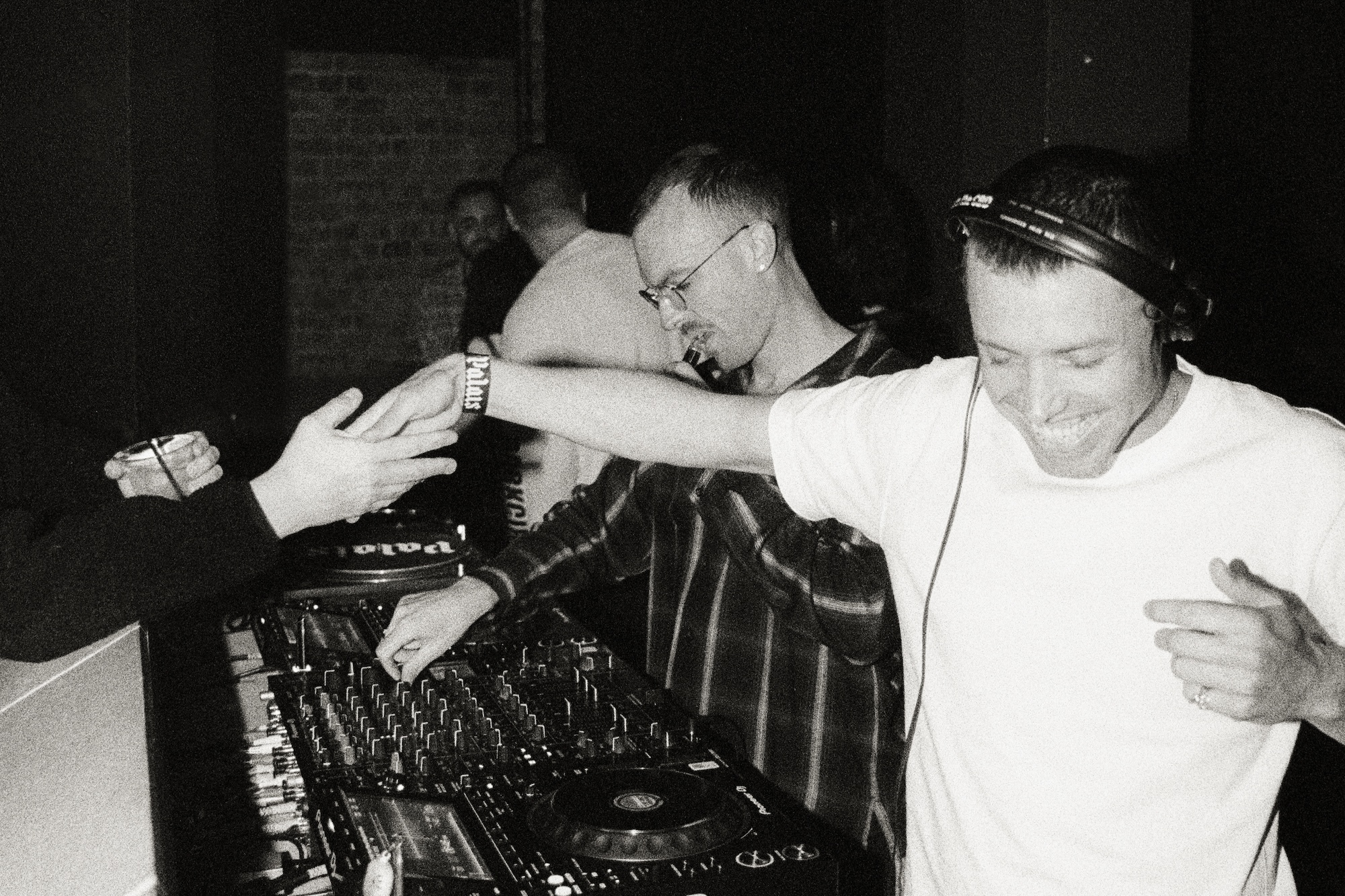 Monochrome image of DJs performing at a club, with mixing decks and a crowd in the background.