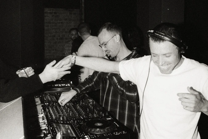 Monochrome image of DJs performing at a club, with mixing decks and a crowd in the background.