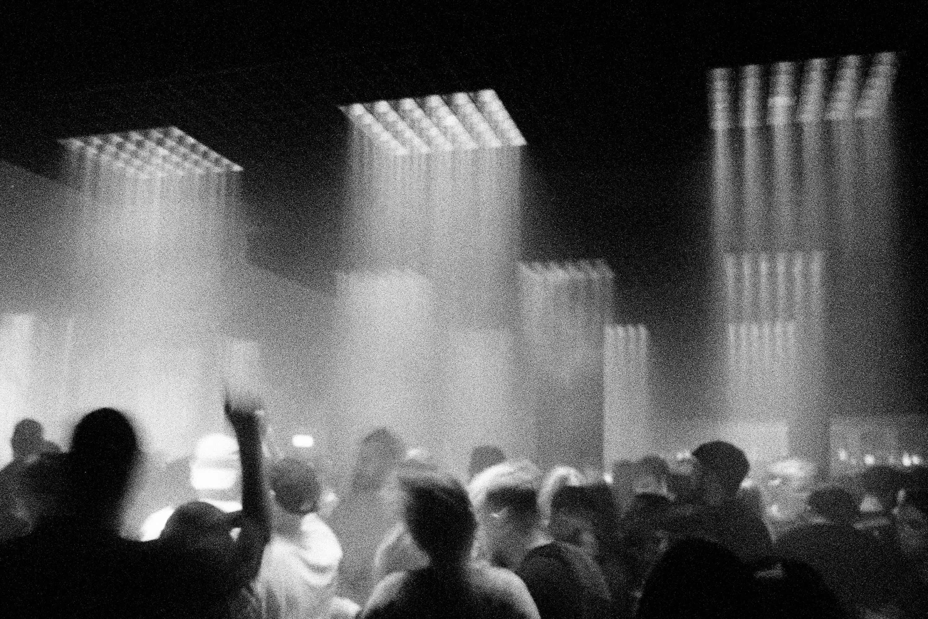 Monochrome shot of a large crowd of people in a dimly lit indoor setting, illuminated by spotlights overhead, creating a vibrant atmosphere.