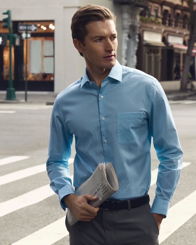 Male model wearing a blue patterned long-sleeve shirt and grey trousers holding a rolled newspaper on a city street.