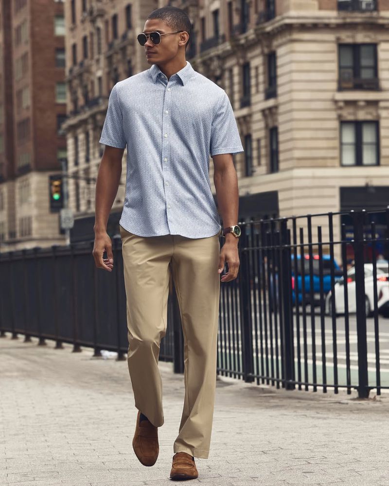 Man wearing sunglasses, short-sleeve patterned blue shirt, khaki trousers and brown shoes walking down a city street
