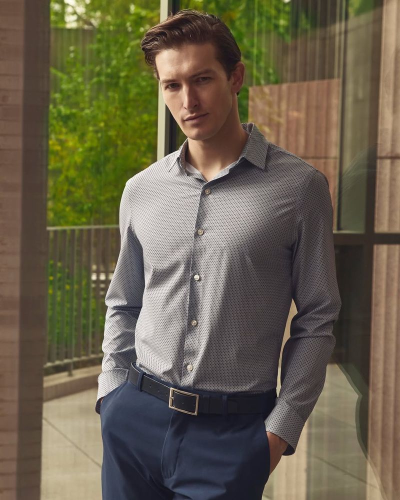 Man wearing a patterned grey button-down shirt and navy blue trousers with a black belt, standing outdoors near a building.