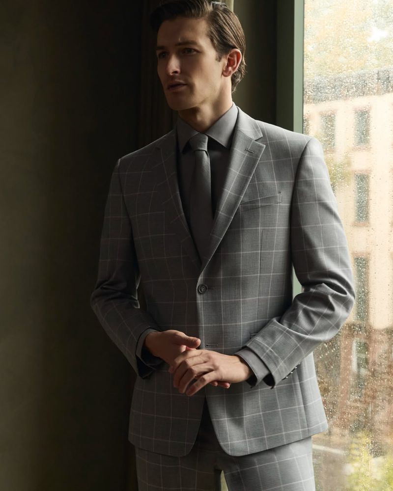 A man in a grey windowpane check suit and tie stands near a window for the Van Heusen Spring Summer 2026 campaign.