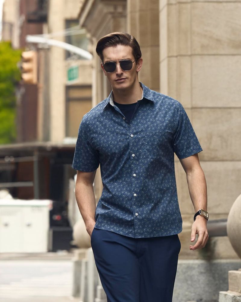 Man wearing sunglasses and a blue short sleeve patterned shirt walking on a city street.
