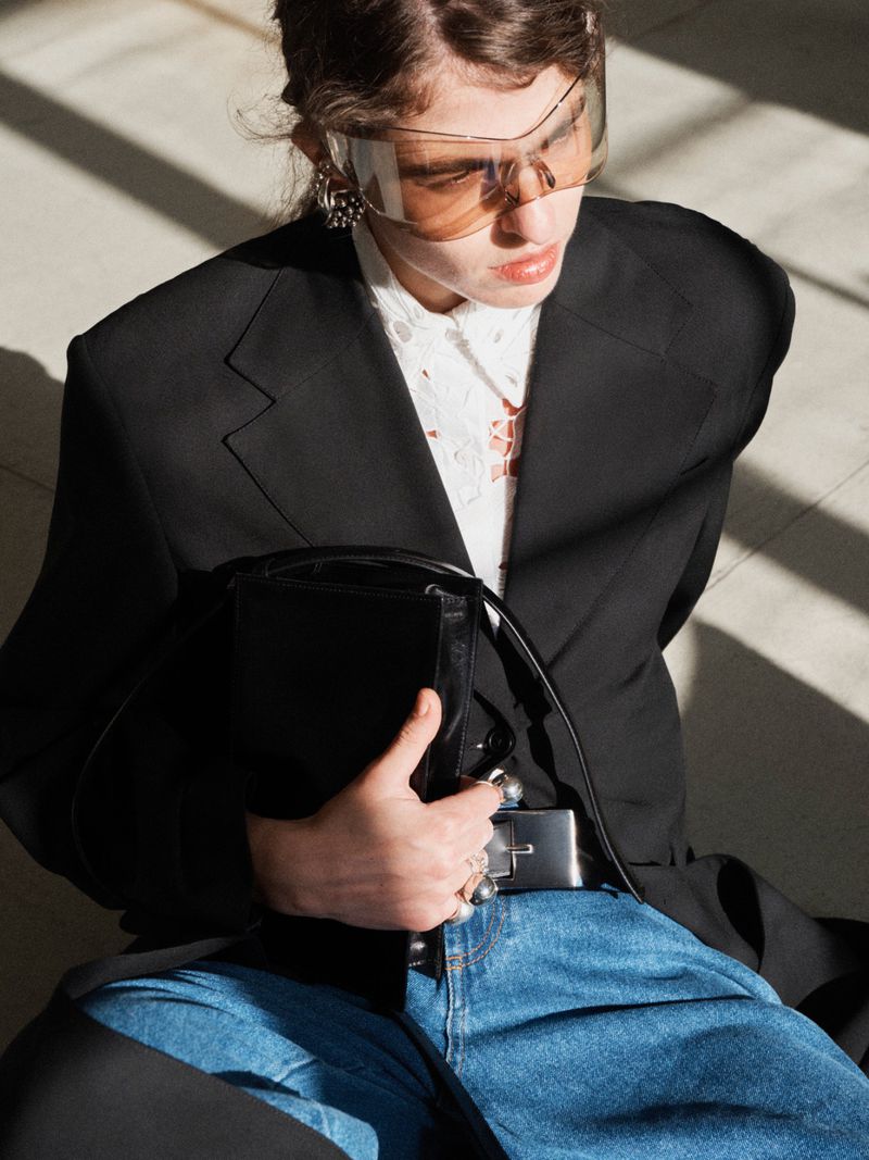 Model wearing a black blazer, white shirt, blue jeans, and visor sunglasses holding a black handbag.