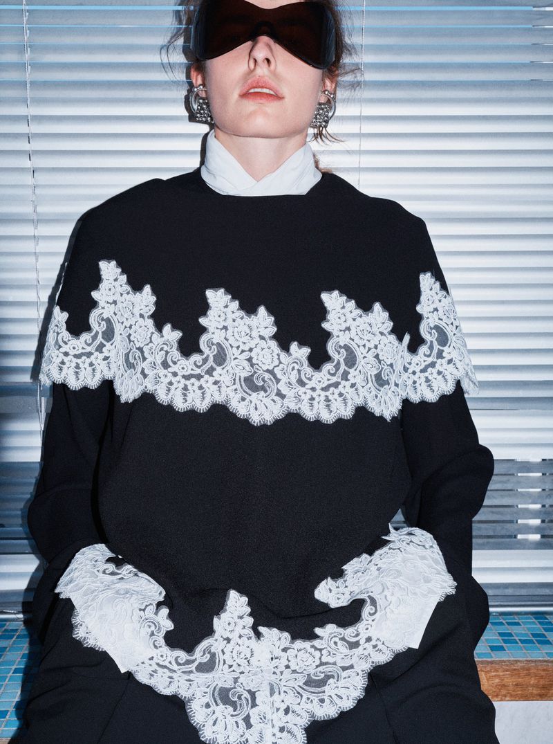 Model wearing dark sunglasses, ornate earrings, and a black garment with white lace trim against horizontal window blinds.
