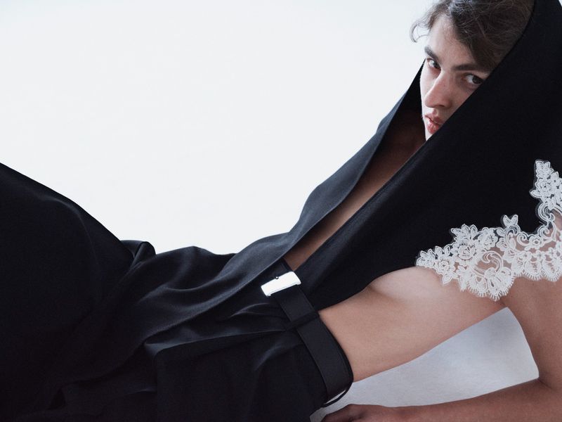 Model in a black hooded garment with white lace trim and a silver buckle belt against a white seamless background