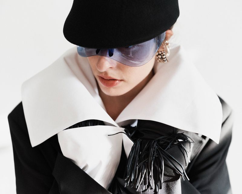 An adult female wears a black hat, sunglasses, and a black jacket with fringe detail, set against a bright white backdrop in a fashion-forward shot.