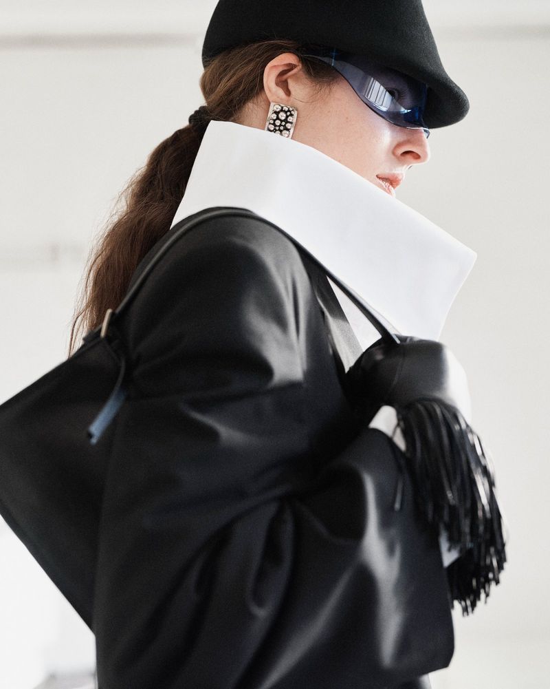 Profile of woman in black hat and sunglasses wearing an oversized jacket with a high white collar and carrying a fringe handbag.