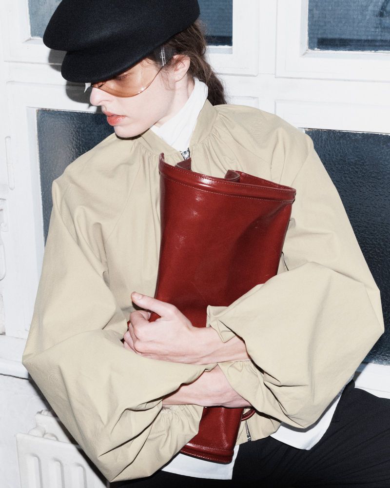 Model wearing a tan jacket, black cap, and amber sunglasses holding a large dark red leather clutch.